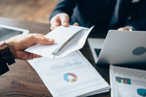 Cropped view of businessman taking envelope with money from investor near papers and laptop on blurred background 
