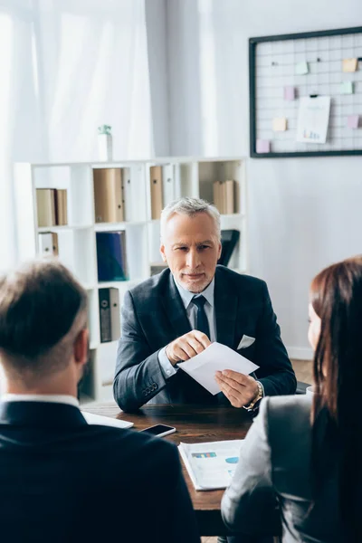 Rijpere Adviseur Houdt Enveloppe Buurt Van Zakelijke Mensen Wazig Voorgrond — Stockfoto