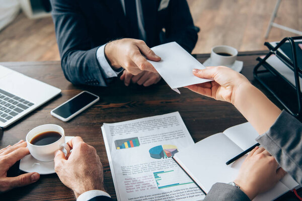 Cropped view of investor giving envelope to businesswoman near documents and cups of coffee on table 