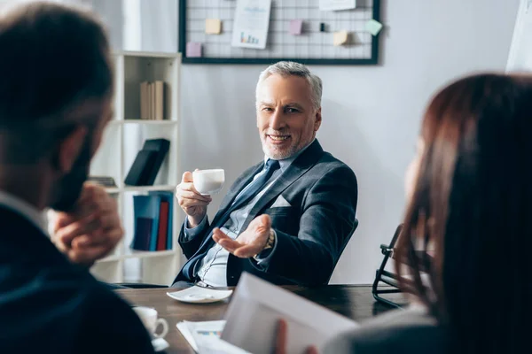 Smiling Investor Coffee Cup Pointing Business People Papers Blurred Foreground — Stock Photo, Image