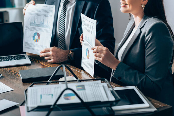 Cropped view of smiling businesswoman holding document with charts near colleague and devices on table 