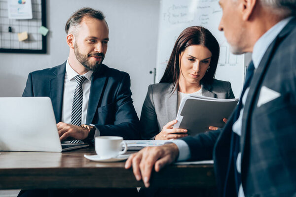 Улыбающийся бизнесмен с ноутбуком сидит рядом с коллегой с бумажной папкой и инвестором на размытом переднем плане в офисе 