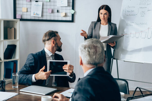 Businessman with digital tablet looking at businesswoman with paper folder near flipchart and investor on blurred foreground 