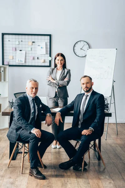 Ofisteki iş ortaklarının yanında kameraya bakan gülümseyen iş kadını 