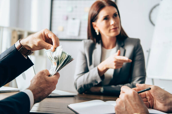 Businessman holding money near colleague and businesswoman pointing with finger on blurred background 