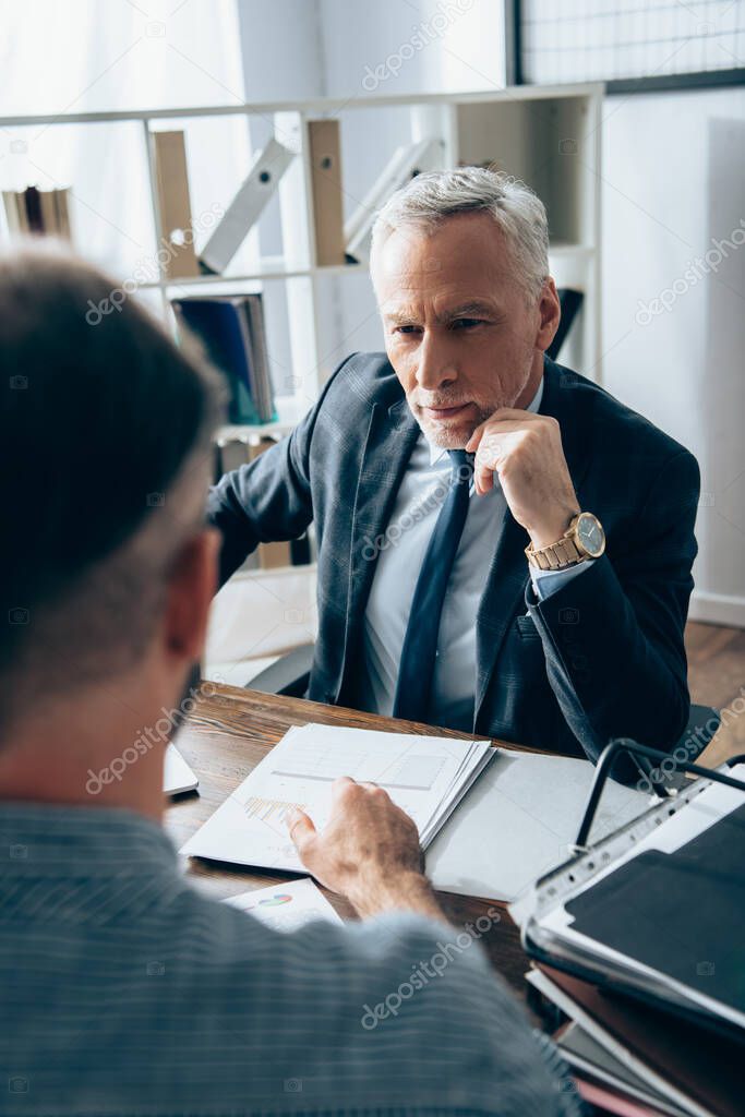 Mature investor looking at businessman on blurred foreground near papers with charts on table in office 