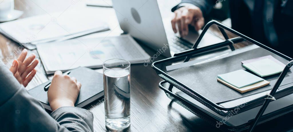 Cropped view of businesswoman holding pen near notebook and glass of water during consultation with investor on blurred background, banner 