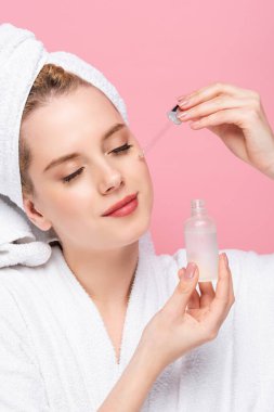 Kafasında havlu olan genç bir kadın pembeye izole edilmiş yüzüne serum uyguluyor.