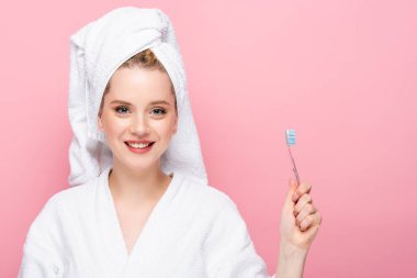 Kafasında havluyla bornozlu gülümseyen genç bir kadın pembe üzerinde diş fırçası tutuyor.