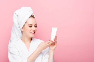 happy young woman in bathrobe with towel on head holding tube with hand cream isolated on pink clipart