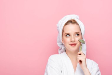 dreamy young woman in bathrobe with towel on head using jade roller isolated on pink clipart