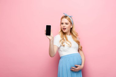 Şok olmuş sarışın hamile kadın pembe arka planda akıllı telefon gösteriyor.