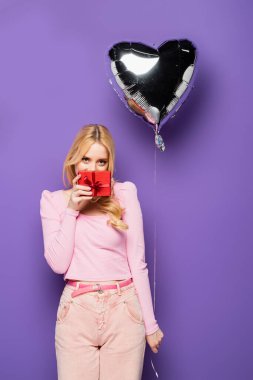Sarışın, genç bir kadın elinde balon şeklinde bir kalp ve mor arka planda hediye tutuyor.