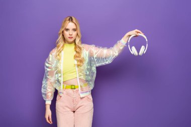 Mor arka planda kulaklık tutan, renkli kıyafetli, üzgün sarışın genç bir kadın.