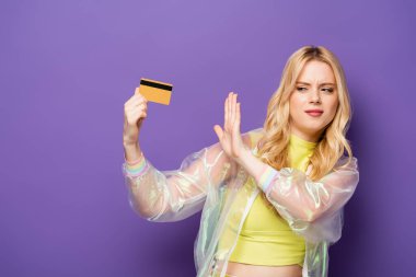 Renkli kıyafetli sarışın genç bir kadın mor arka planda kredi kartını reddediyor.
