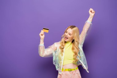 Mor arka planda kredi kartı gösteren renkli elbiseli mutlu sarışın kadın.