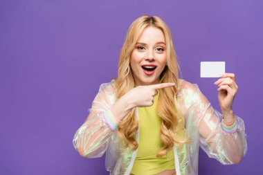 shocked blonde young woman in colorful outfit pointing at blank card on purple background clipart