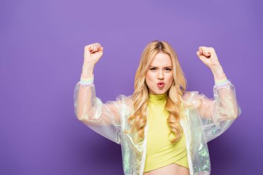 blonde young woman in colorful outfit showing yeah gesture on purple background clipart