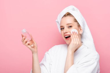 happy young woman in bathrobe with towel on head cleansing face with micellar water and cotton pad isolated on pink clipart