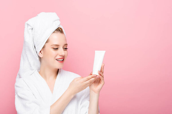 happy young woman in bathrobe with towel on head holding tube with hand cream isolated on pink