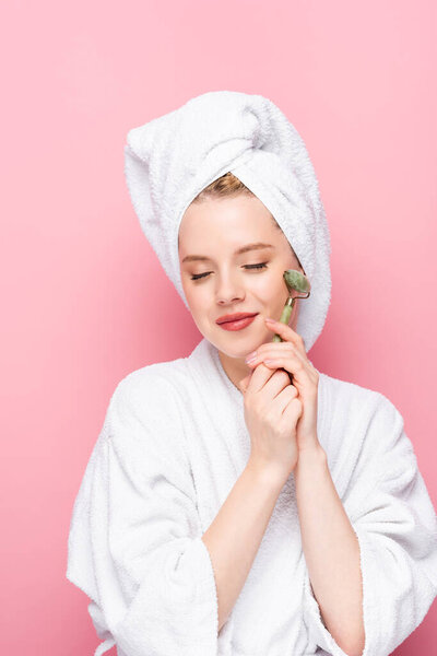 молодая женщина в халате с полотенцем на голове с нефритовым роликом с закрытыми глазами изолированы на розовый