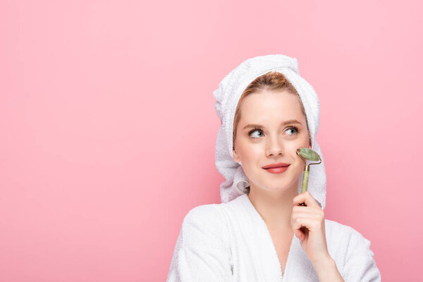 dreamy young woman in bathrobe with towel on head using jade roller isolated on pink