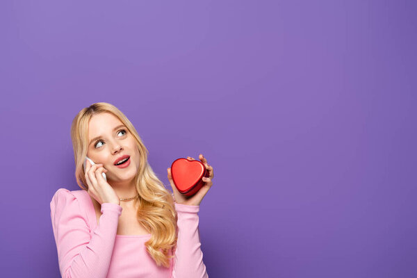 blonde young woman with red heart shaped box talking on smartphone on purple background