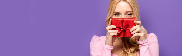 blonde young woman holding red gift box near face on purple background, banner