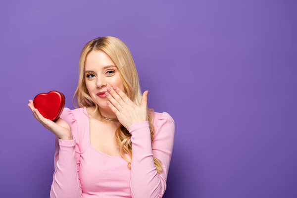 shy blonde young woman holding red heart shaped box on purple background