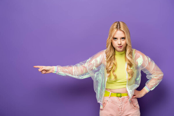 angry blonde young woman in colorful outfit pointing aside on purple background