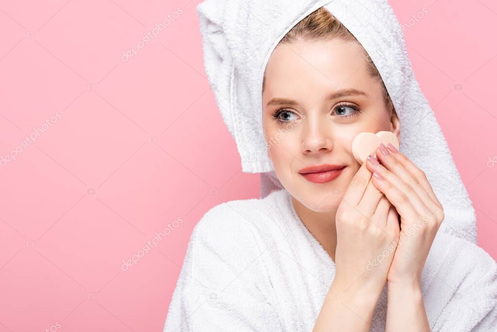 young woman in bathrobe with towel on head using facial heart shaped sponge isolated on pink