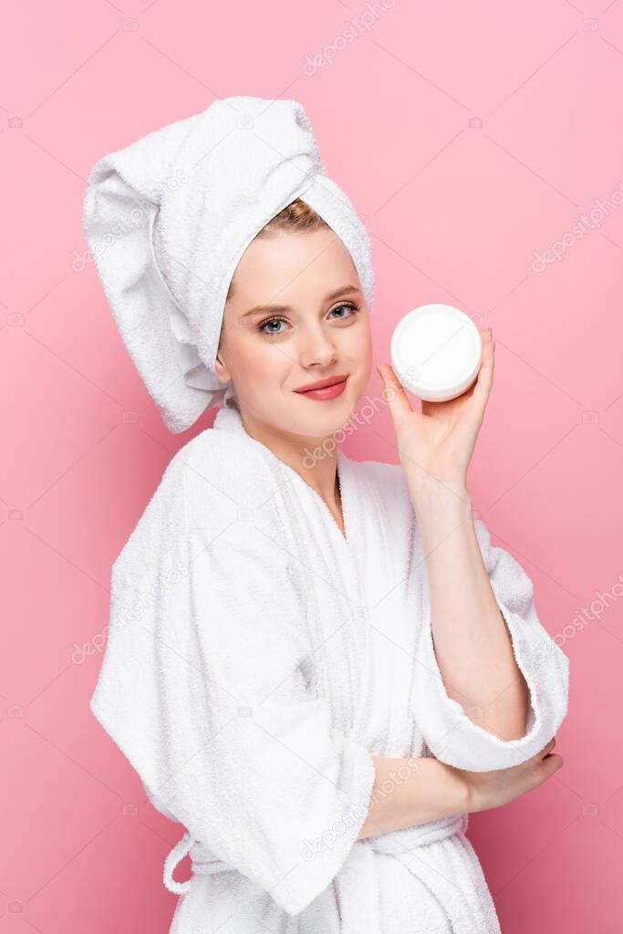 young woman in bathrobe with towel on head holding cosmetic cream isolated on pink