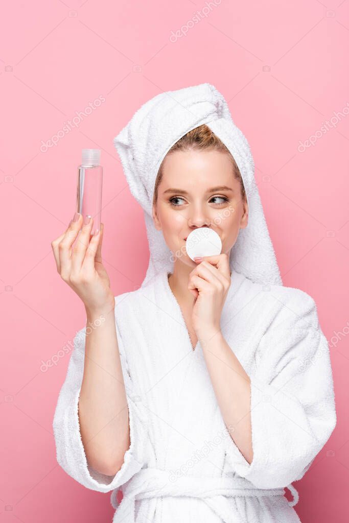 young woman in bathrobe with towel on head with micellar water and cotton pad isolated on pink