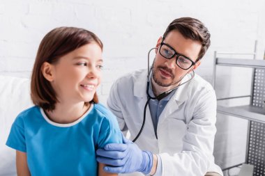 pediatrician examining smiling girl with stethoscope in hospital, blurred foreground