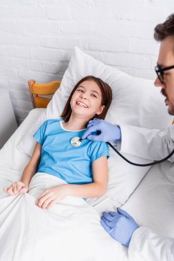 pediatrician examining cheerful girl with stethoscope in hospital, blurred foreground