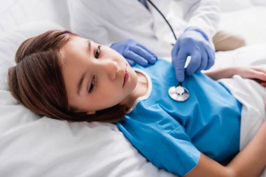 pediatrician examining diseased girl with stethoscope, blurred background