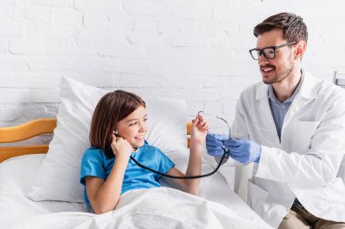 Neşeli çocuk doktoru hastane yatağında steteskopu mutlu kıza gösteriyor.