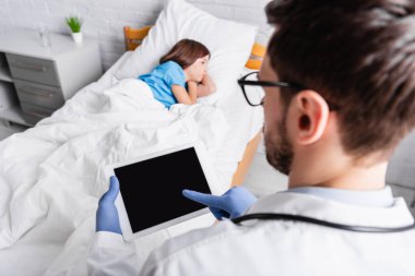 pediatrician using digital tablet near sick girl lying in bed, blurred foreground