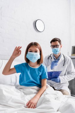 Tıbbi maskeli çocuk bulanık arka planda panosu olan doktorun yanında iyi bir hareket gösteriyor.