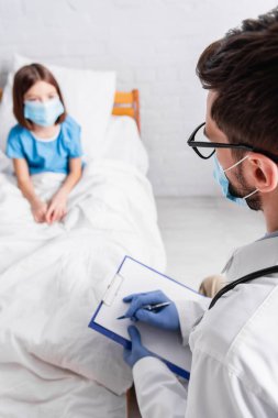 Pediyatrist, bulanık arka planda, tıbbi maskeli hasta bir kızın yanında, panoya teşhis yazıyor.