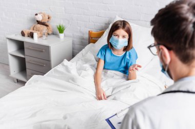 Tıbbi maskeli çocuk ön planda bulanık bir şekilde doktorla konuşurken elini gösteriyor.