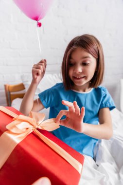 smiling girl opening birthday gift while holding festive balloon in hospital, blurred background