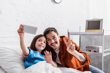 happy man showing victory gesture near daughter waving hand while taking selfie on smartphone, blurred foreground