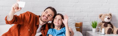 happy girl waving hand near dad taking selfie on mobile phone in hospital, banner