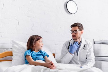 Çocuk doktoru yatakta bir kızla konuşurken boğazını işaret ediyor.