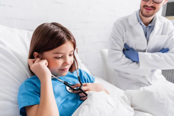 微笑的女孩使用听诊器接近微笑的医生在模糊的背景下 — 图库照片