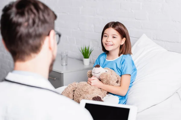 Szczęśliwa Dziewczyna Misiem Patrząc Pediatrę Tabletem Cyfrowym Niewyraźnym Pierwszym Planie — Zdjęcie stockowe