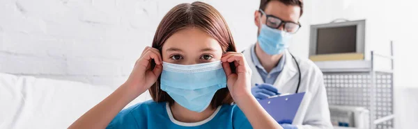 sick kid fixing medical mask while looking at camera near doctor writing diagnosis on blurred background, banner