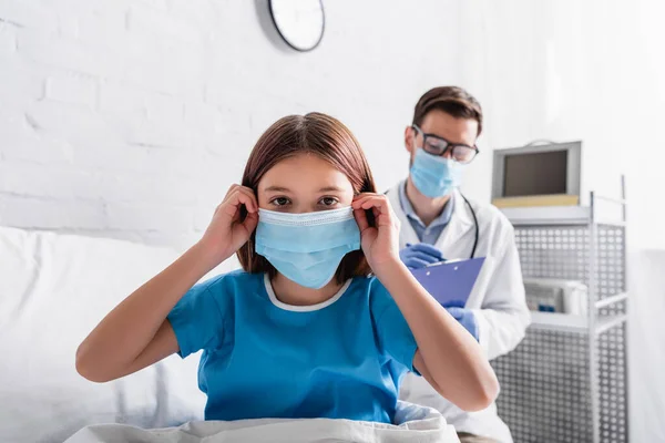 Diseased Girl Looking Camera While Fixing Medical Mask Doctor Writing — Stock Photo, Image
