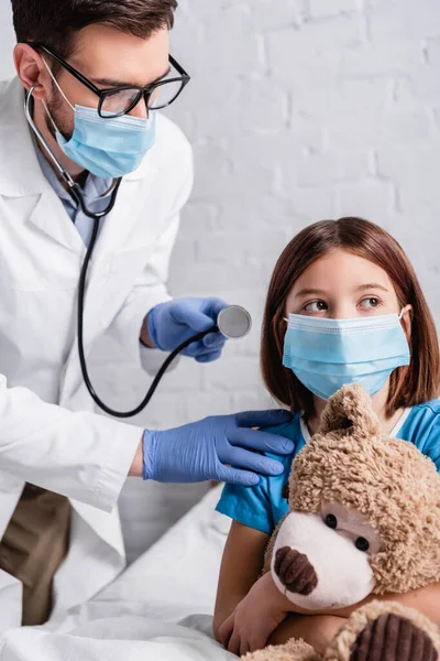 Παιδίατρος Στηθοσκόπιο Κοντά Κορίτσι Ιατρική Μάσκα Κάθεται Στο Κρεβάτι Αρκουδάκι — Φωτογραφία Αρχείου